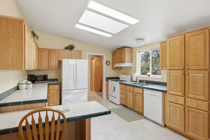 Kitchen featuring light wood cabinetry, white appliances, skylight, and durable tile countertops with green trim