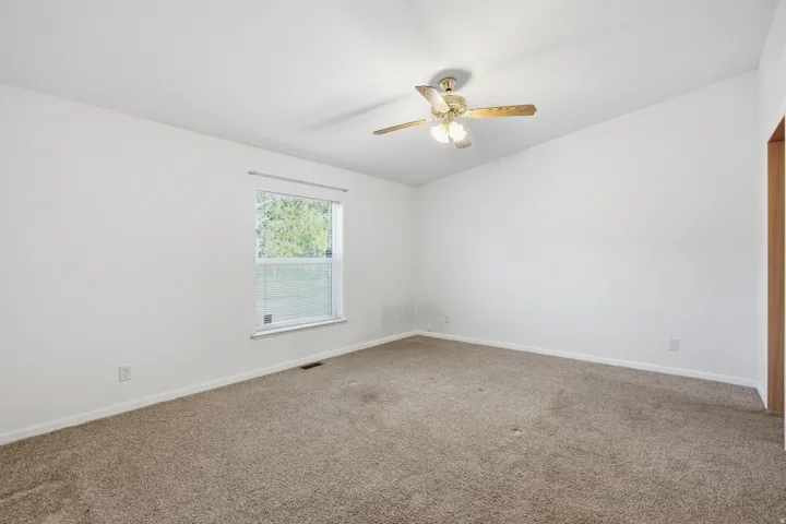 Carpeted room featuring a ceiling fan with light fixture, a window with exterior tree scenery, and white wall finishes