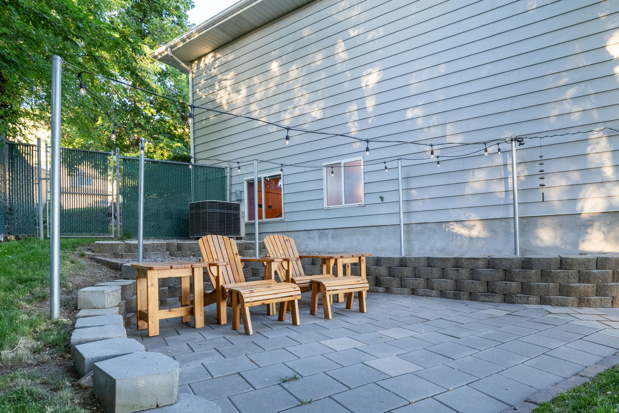 Paved patio area with string lighting, bordered by a concrete block retaining wall and a chain-link fence