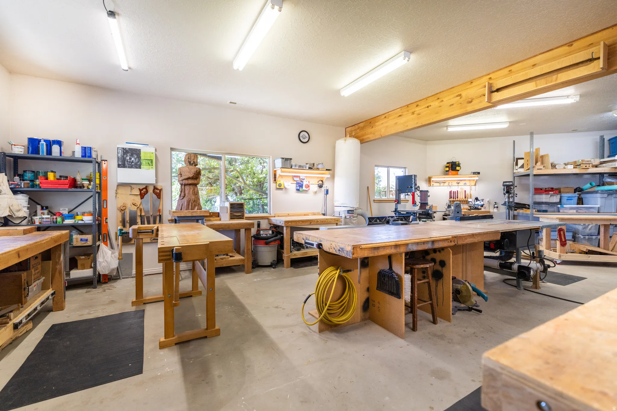 Spacious workshop featuring concrete flooring, overhead fluorescent lighting, exposed wood beams, multiple large workbenches, and natural light from a prominent window