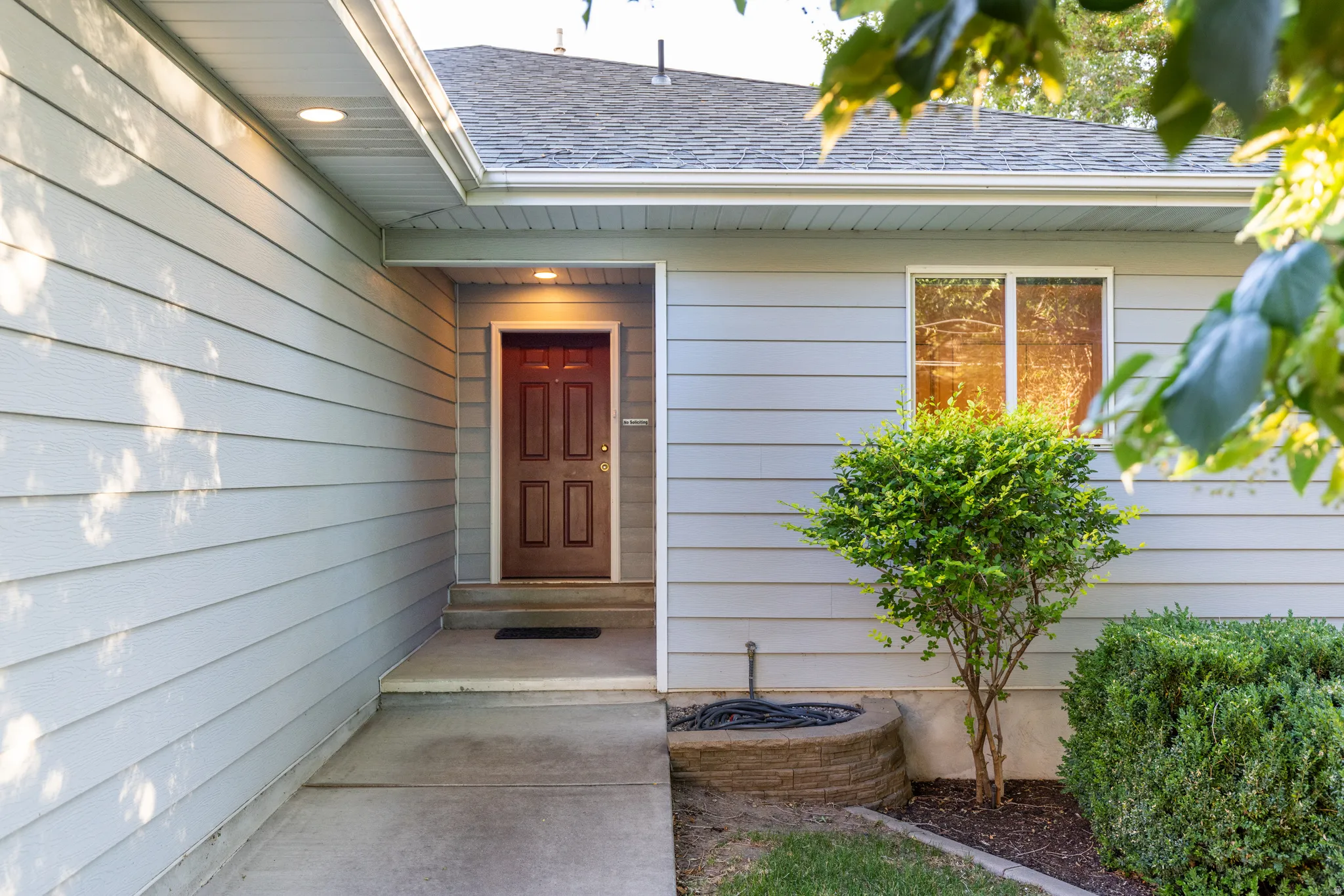Front entrance featuring a paneled red door, light blue siding, a concrete pathway, a shingled roof, and perimeter landscaping