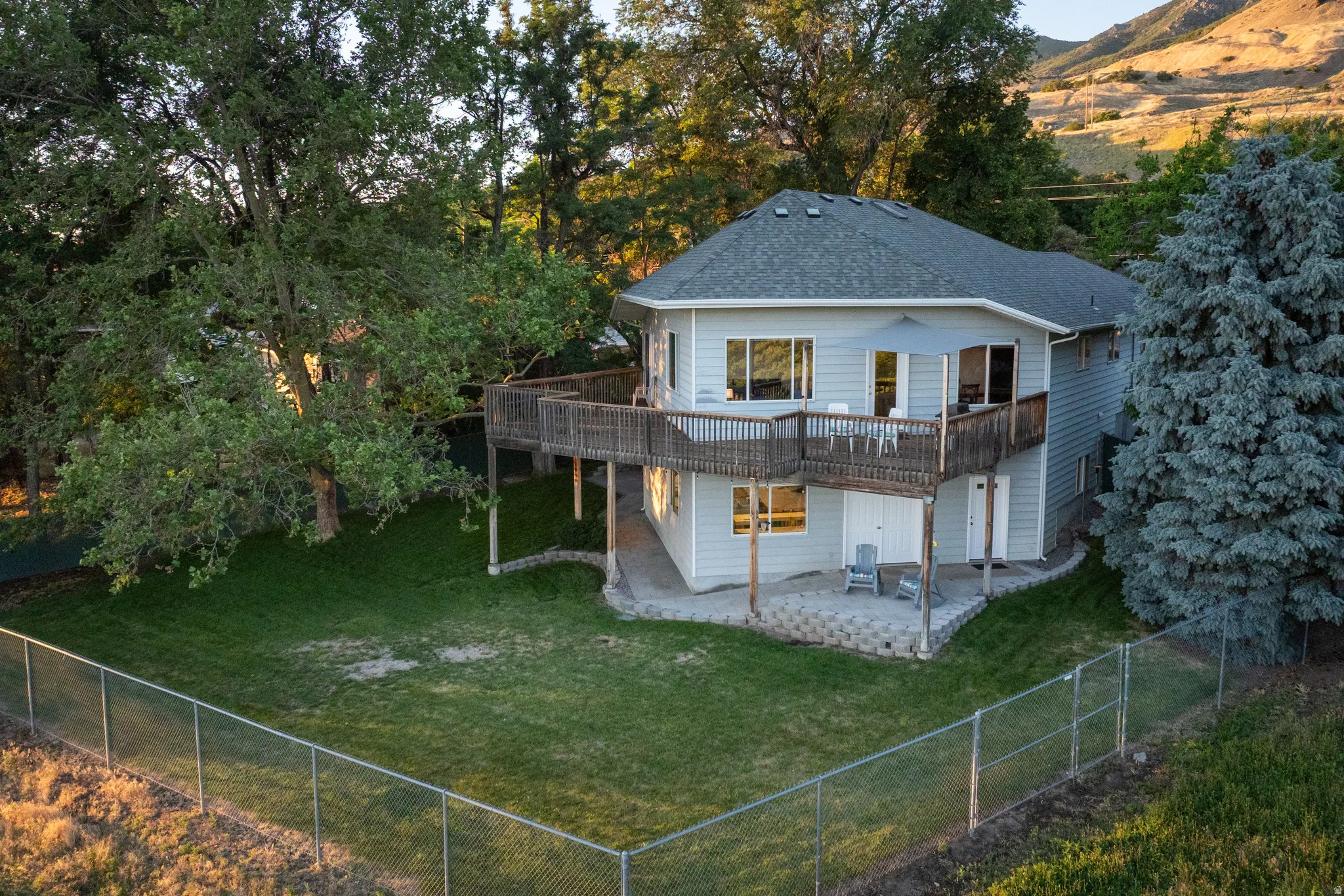 Light blue siding residence featuring a spacious wood-finish deck with railing, a lower-level patio with stone pavers, and a large grassy yard enclosed by chain link fencing