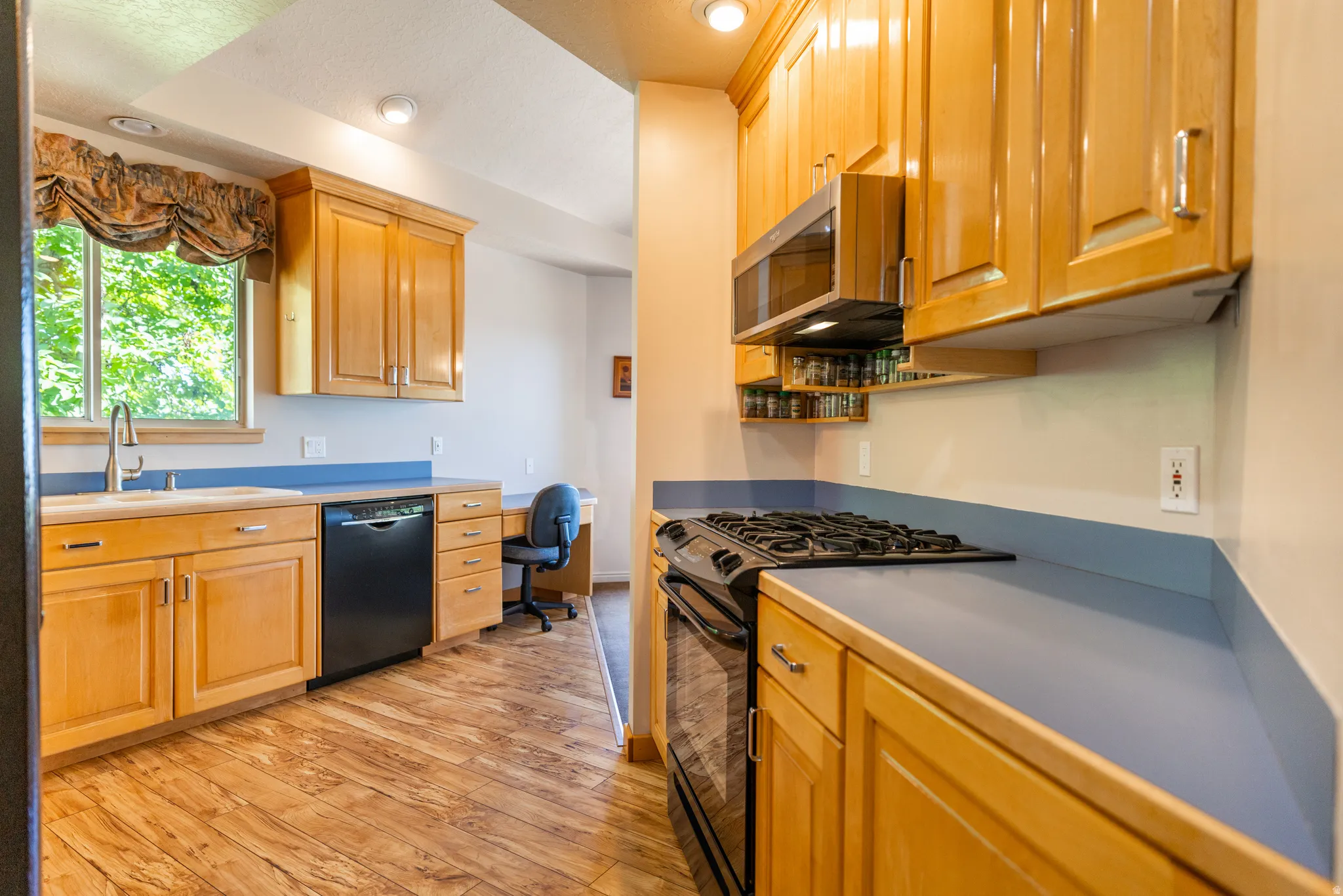Kitchen featuring light wood cabinetry, a black gas range, built-in microwave, wood-finish flooring, and a window with a stainless steel sink