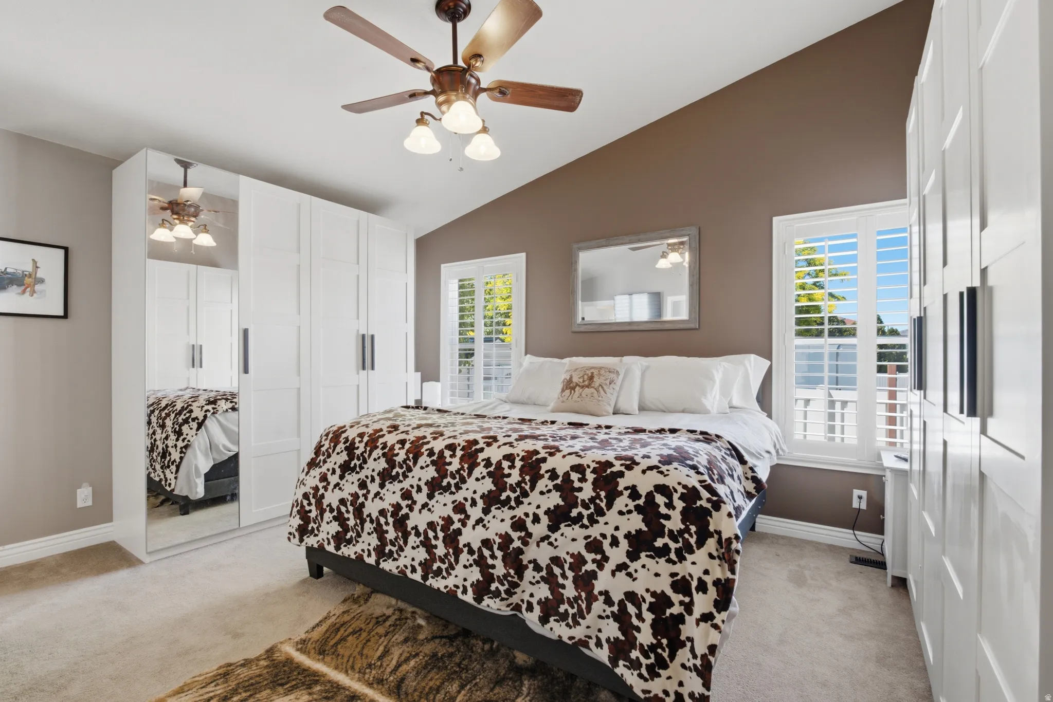 Vaulted ceiling bedroom featuring white plantation shutters, a wood-finish ceiling fan, mirrored closet doors, and light neutral carpeting