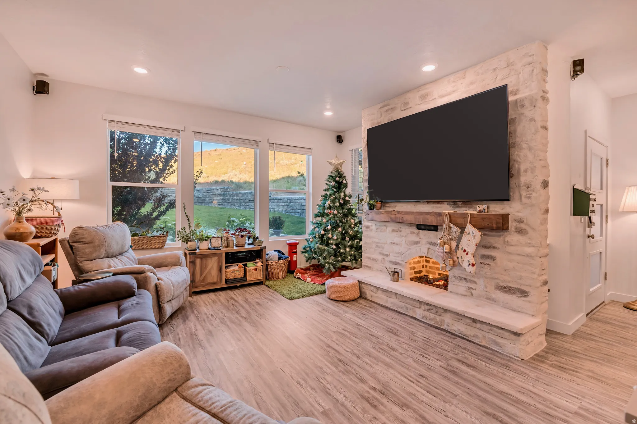 Spacious living area featuring expansive windows, wood-finish flooring, a stone accent wall, and a rustic wood mantel
