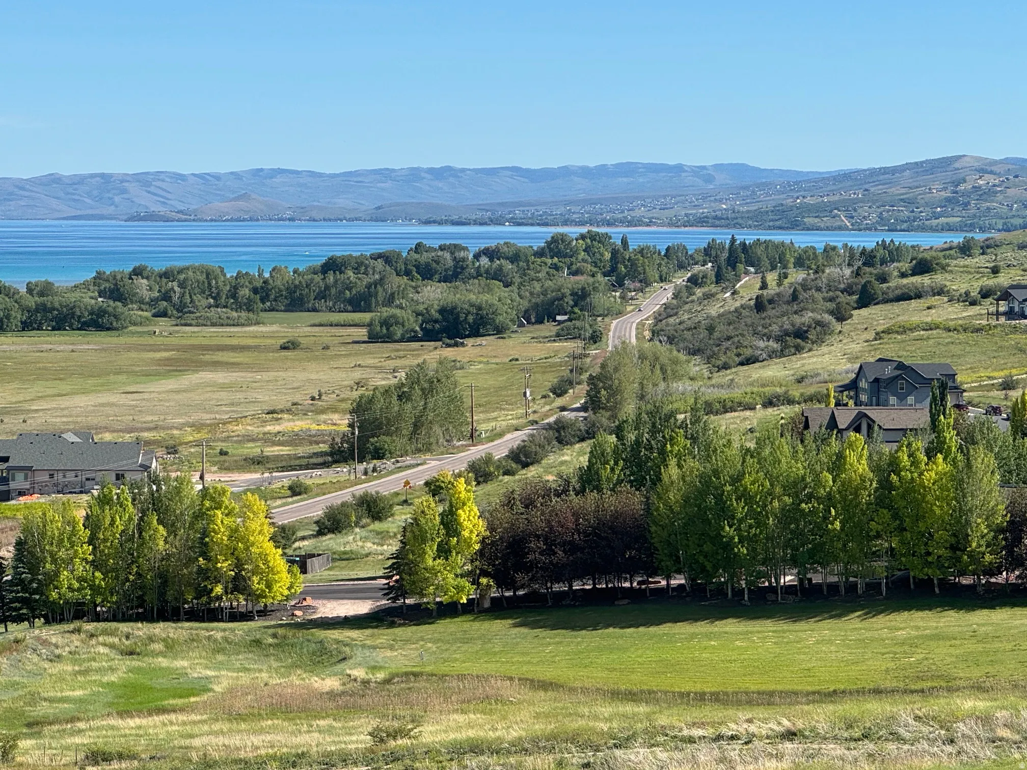 Expansive landscape featuring a large body of water, distant mountains, a winding road, and scattered residential properties