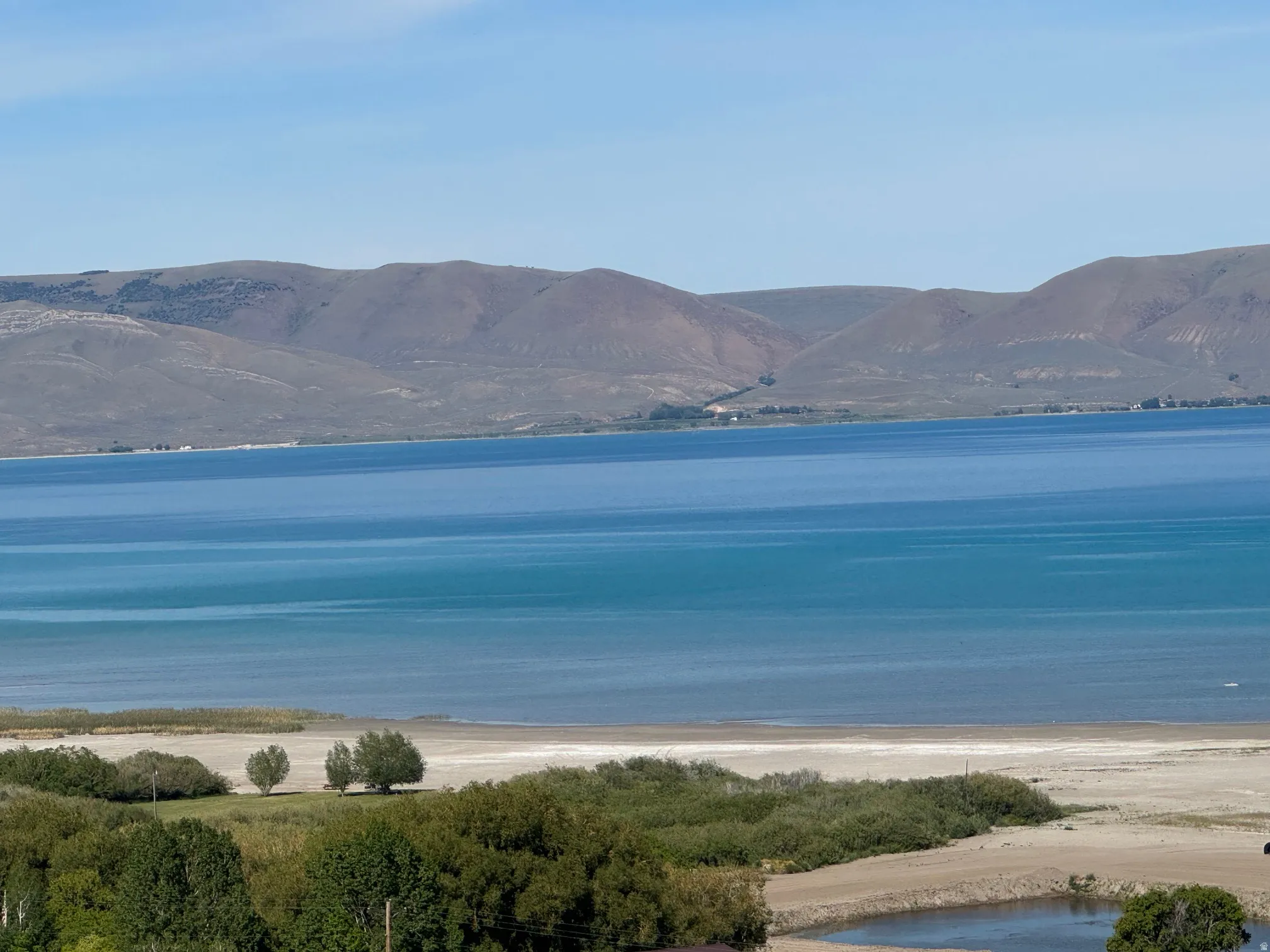Expansive water frontage with a sandy shore, bordered by mature trees and a tranquil pond, against a backdrop of distant hills