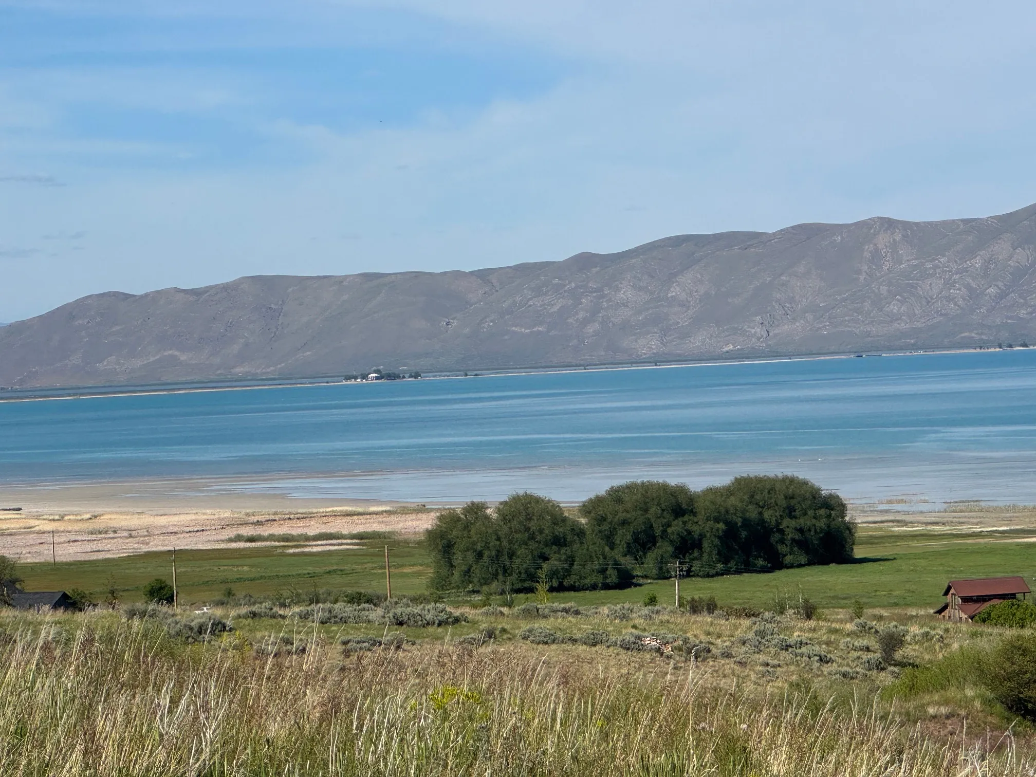 Expansive water body with a mountainous backdrop, featuring a distant landmass, foreground vegetation, and a small structure with a reddish roof