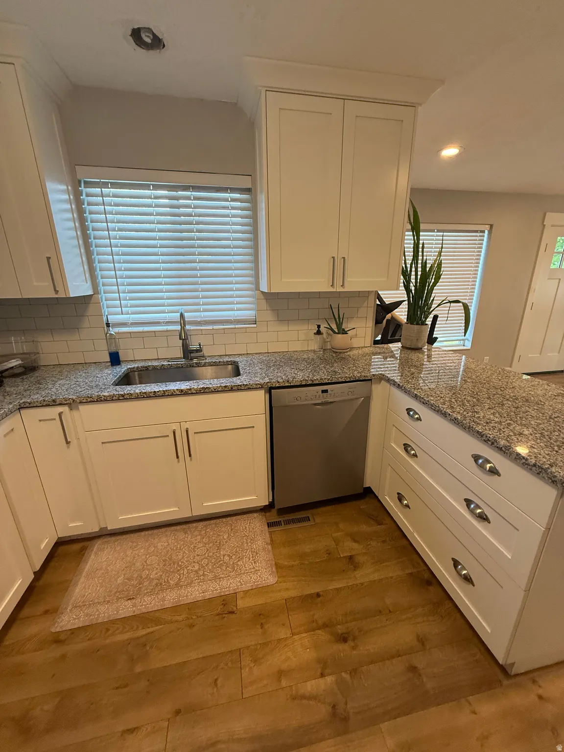 White shaker-style cabinetry, granite countertops, subway tile backsplash, stainless steel dishwasher, and wood-finish flooring