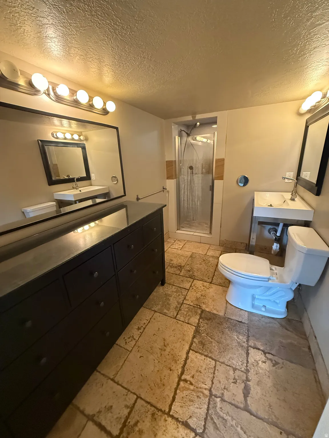 Stone tile flooring, glass-enclosed shower, wall-mounted sinks with chrome fixtures, and vanity lighting