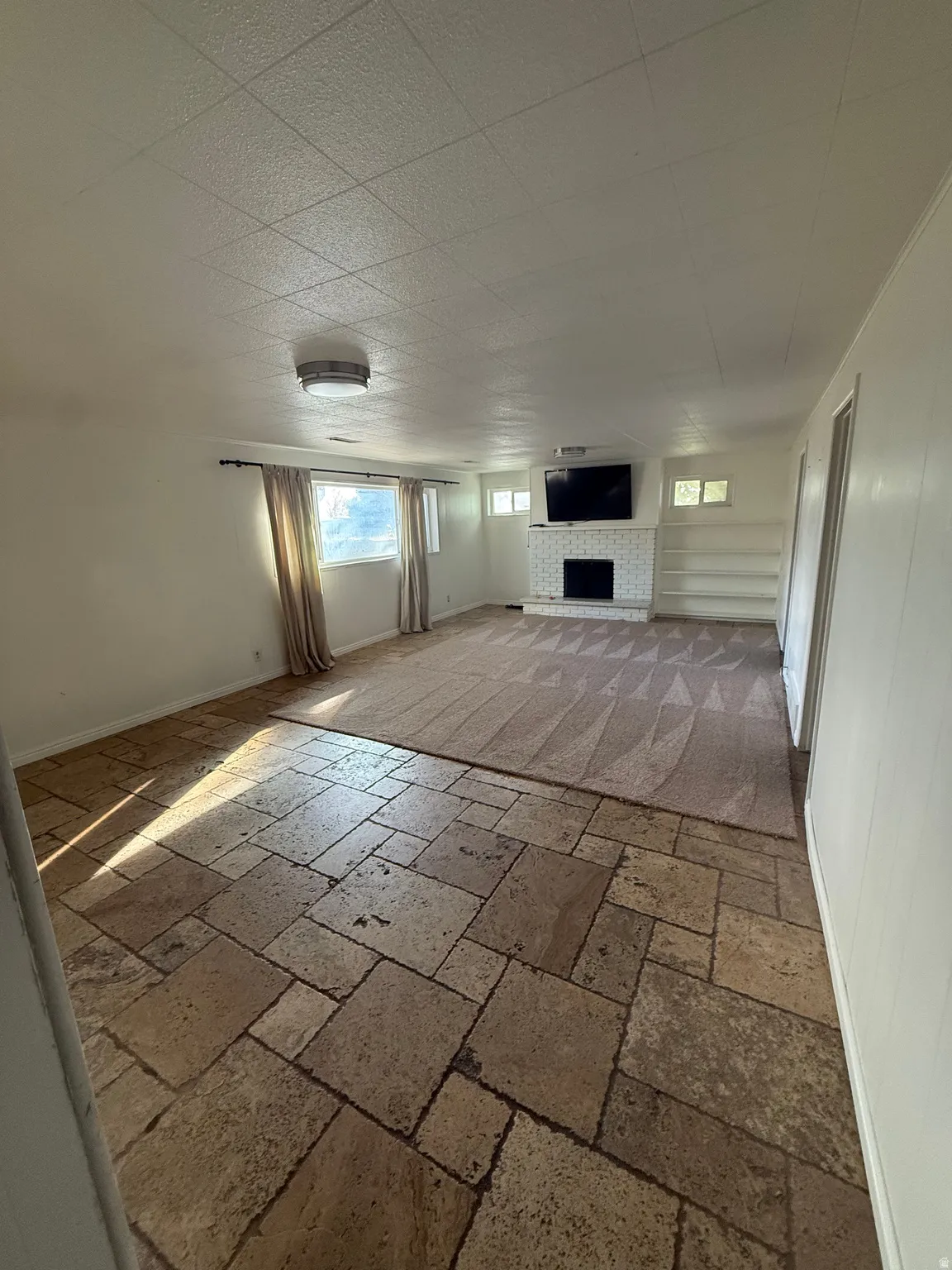 Stone tile flooring transitioning to patterned carpeting, a fireplace with a white brick surround, built-in shelving, and a ceiling-mounted light fixture