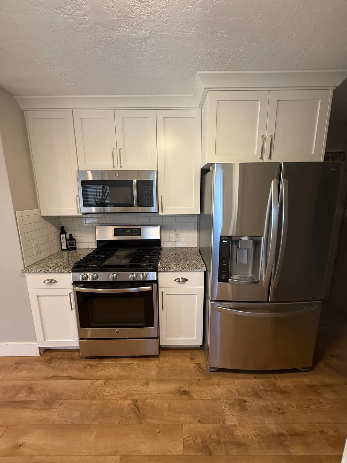 Kitchen featuring white shaker cabinetry with crown molding, stainless steel appliances, speckled stone countertops, a subway tile backsplash, and wood-finish flooring