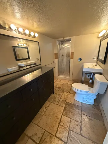 Stone tile flooring, glass-enclosed shower, wall-mounted sinks with chrome fixtures, and vanity lighting