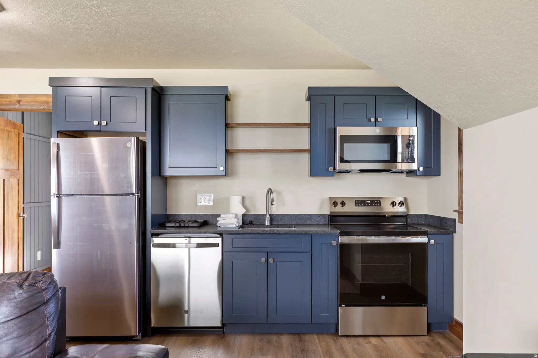 Compact kitchen featuring full-size stainless steel appliances, blue cabinetry, dark countertops, and wood-finish flooring