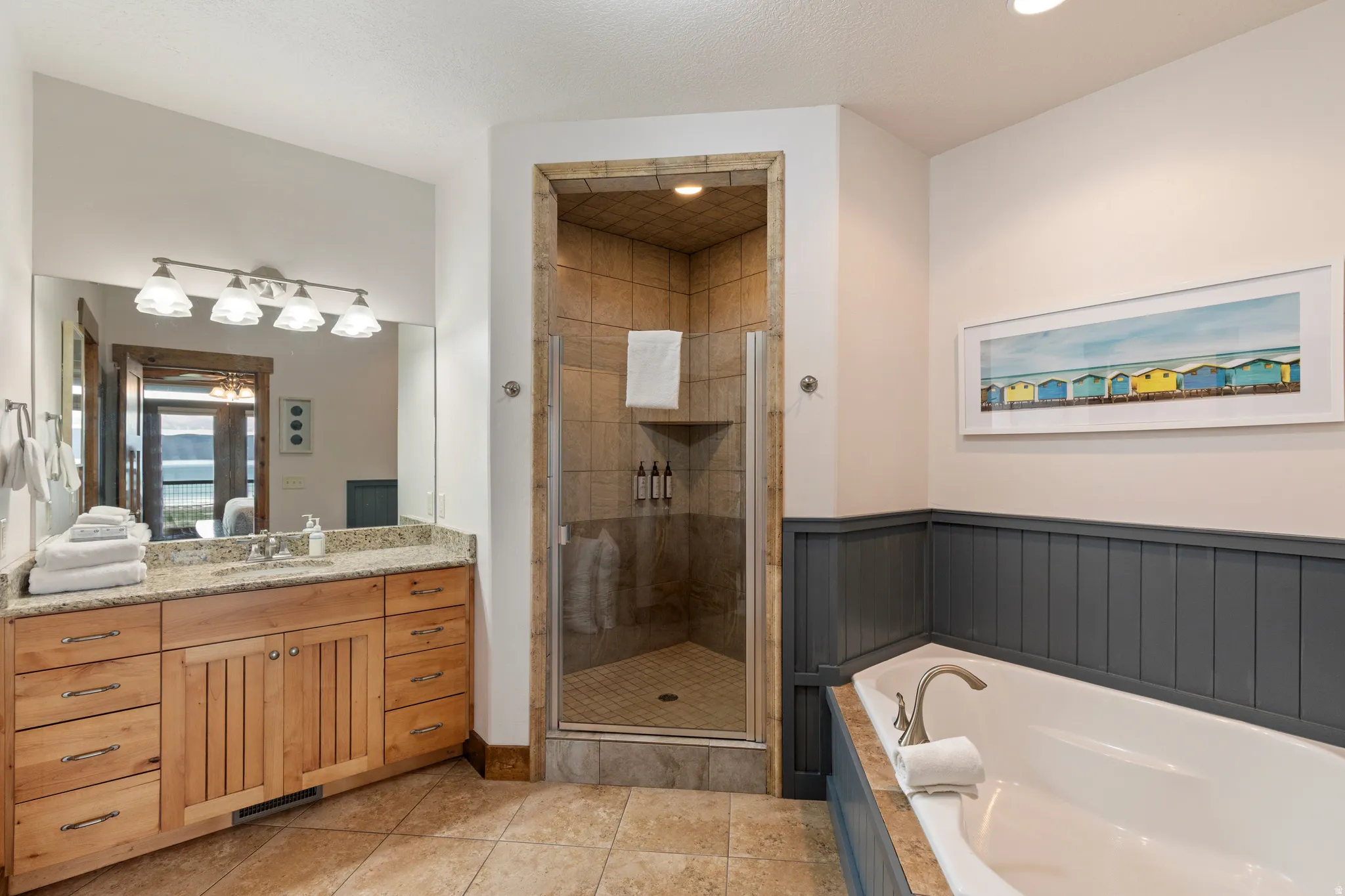 Bathroom featuring a built-in soaking tub, a stand-up shower with glass enclosure, a vanity with a light wood-finish cabinet and stone countertop, tile flooring, and wainscoting