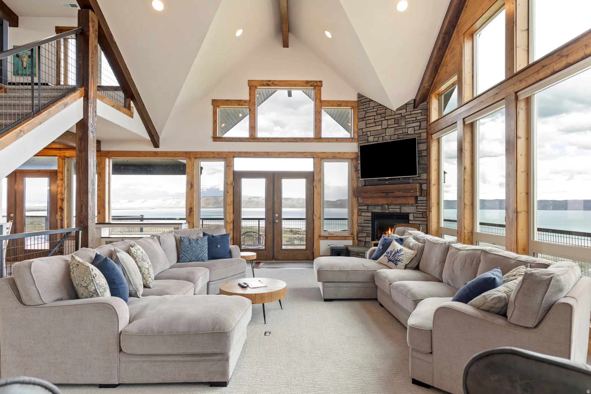 Living area featuring a vaulted ceiling with exposed wood beams, a stone-faced fireplace, a wood-framed full-height window wall, double French doors, and a staircase with metal railings