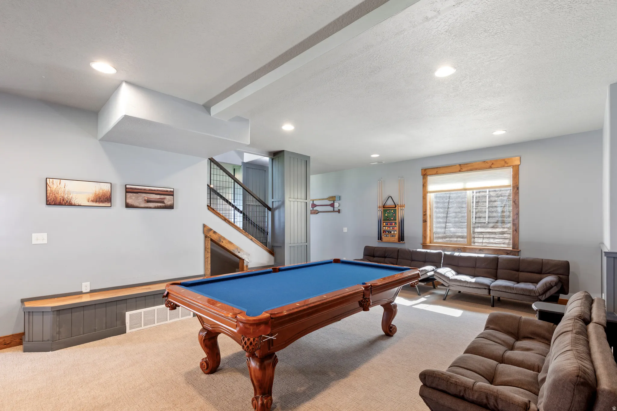 Spacious recreational room featuring recessed lighting, light-toned carpeting, wood-trimmed window, a built-in bench with wood-finish top, and a staircase with black metal railings