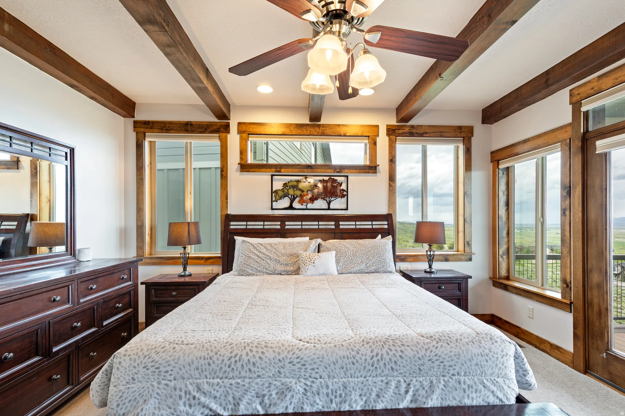 Spacious bedroom featuring exposed wood beams, a ceiling fan with integrated lighting, and multiple windows with wood trim