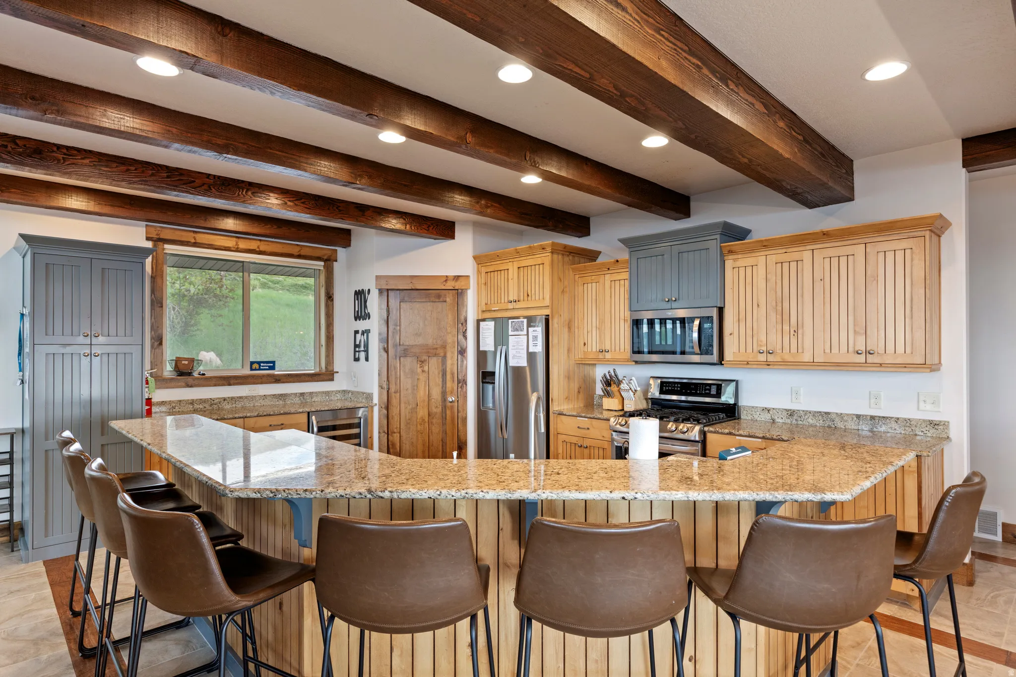 Kitchen featuring a large breakfast bar with stone countertop, exposed wood ceiling beams, wood-finish cabinetry, stainless steel appliances, and built-in recessed lighting
