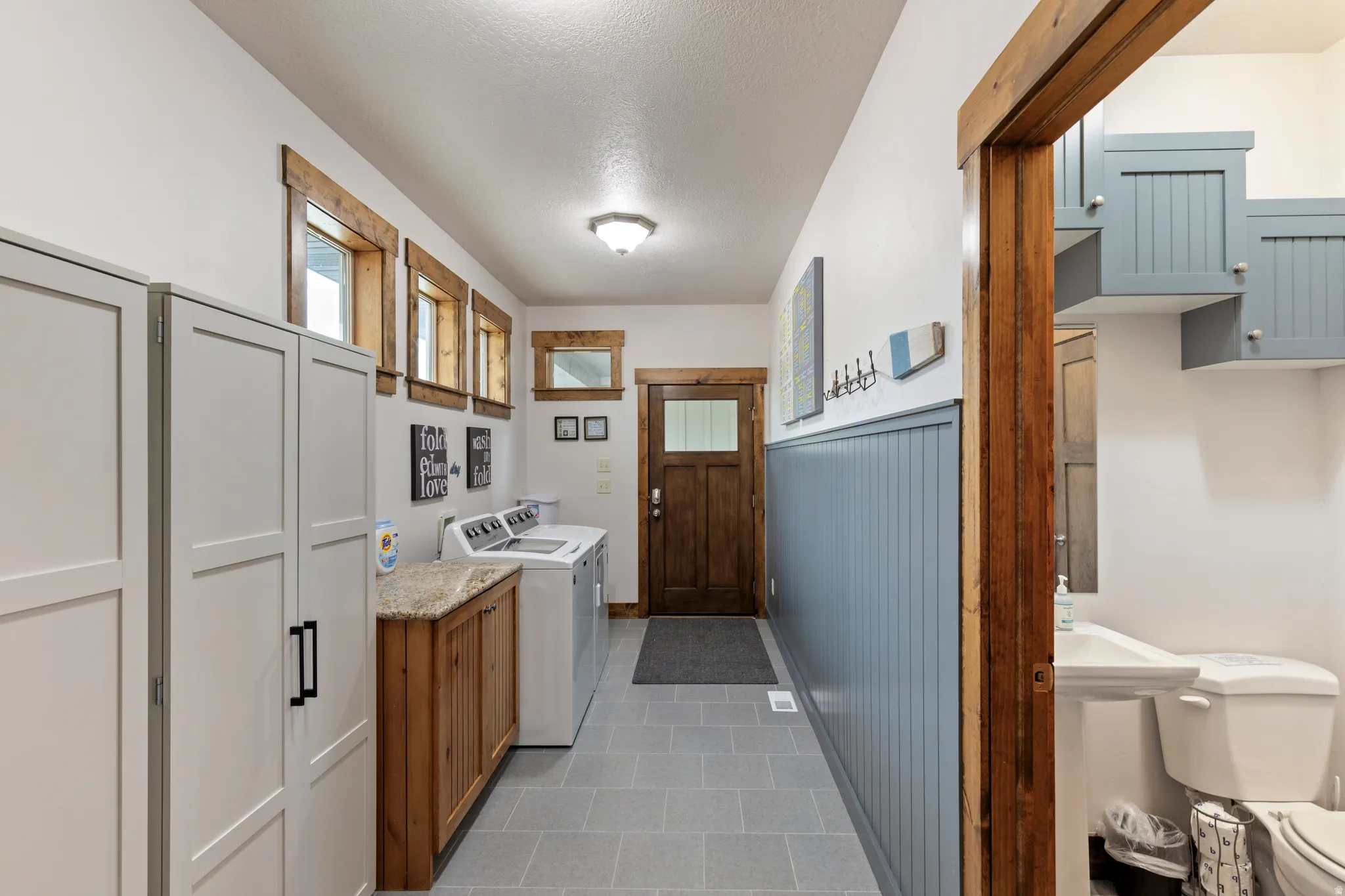 Laundry area with light gray square tile flooring, wood-finish base cabinets with a light-toned countertop, and built-in white paneled storage cabinetry