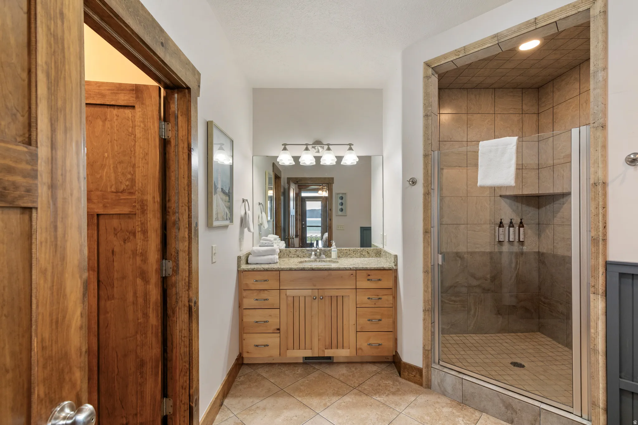 Spacious bathroom featuring a glass-enclosed shower with tile surround, a wood vanity with a stone-finish countertop, and decorative wall-mounted lighting