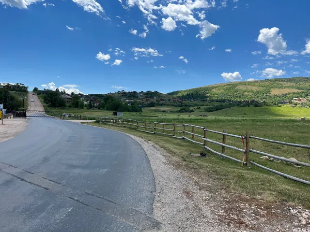 Paved road providing access to hillside properties, bordered by a split-rail fence and expansive green fields