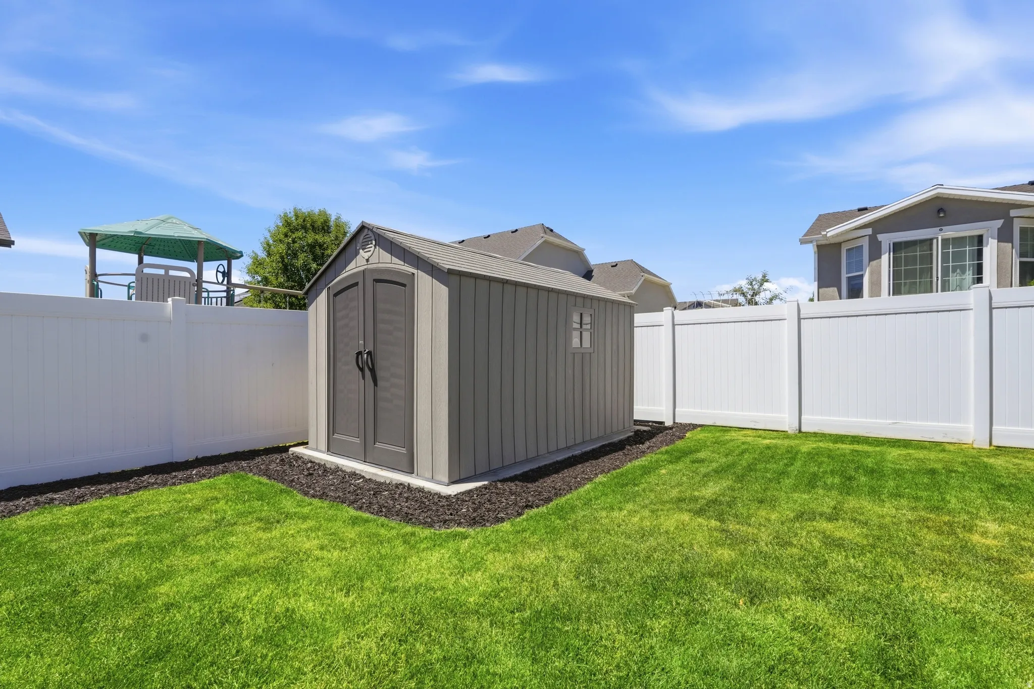 Manicured lawn with a privacy fence, featuring a storage shed with double doors and a window, set on a dark mulch bed