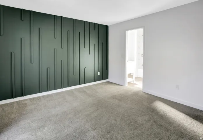 Accent wall featuring vertical paneling, light gray carpeting, white baseboards, and a doorway leading to a bright bathroom with a white vanity