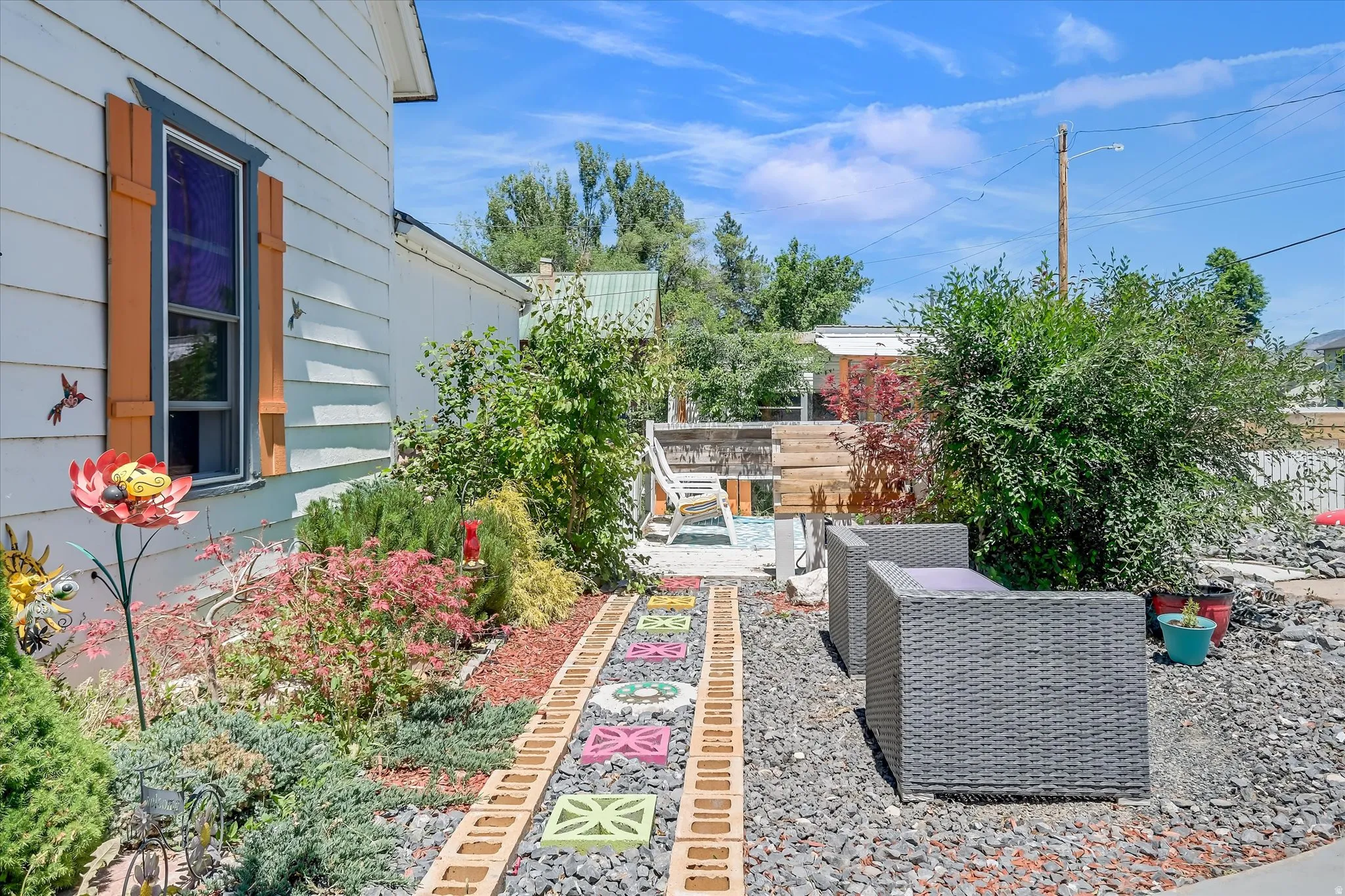 Exterior siding with orange-toned window shutters, a decorative garden pathway, mature landscaping, and an outdoor seating area