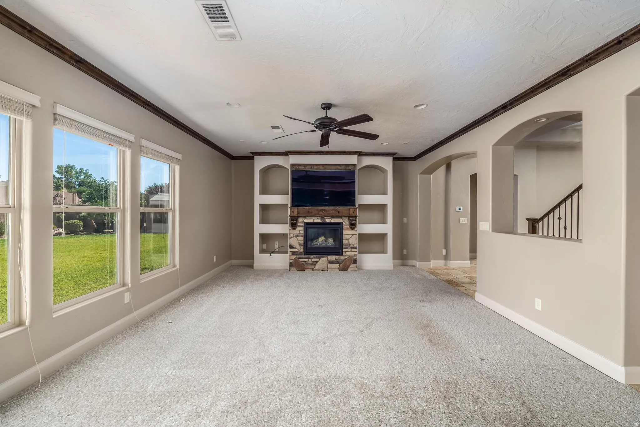Spacious living area featuring a stone-clad fireplace with built-in shelving, wood-finish crown molding, and multiple windows