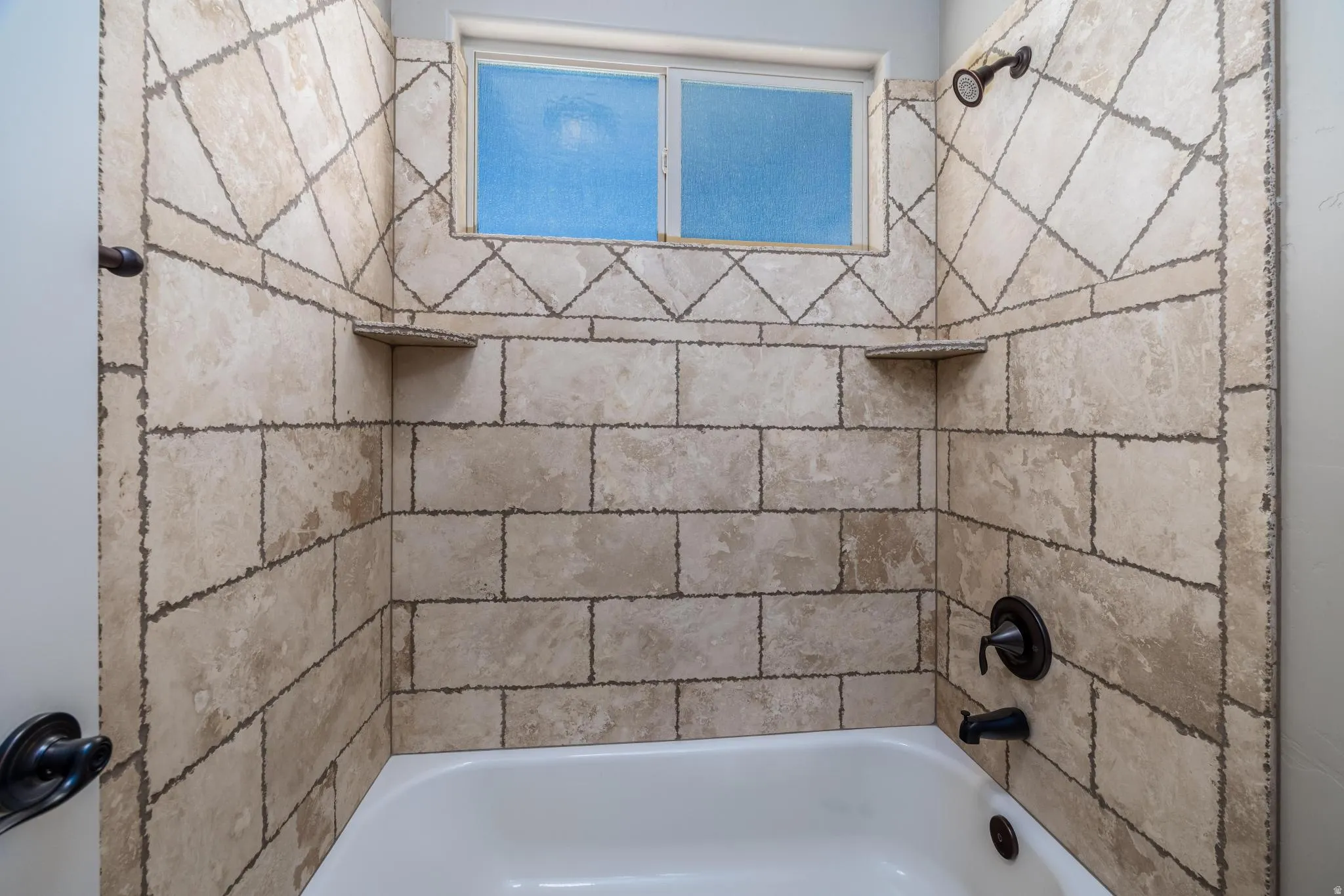 Bathtub and shower combination featuring neutral-toned tile surround with decorative accents, built-in corner shelves, a window with obscure glass, and oil-rubbed bronze fixtures