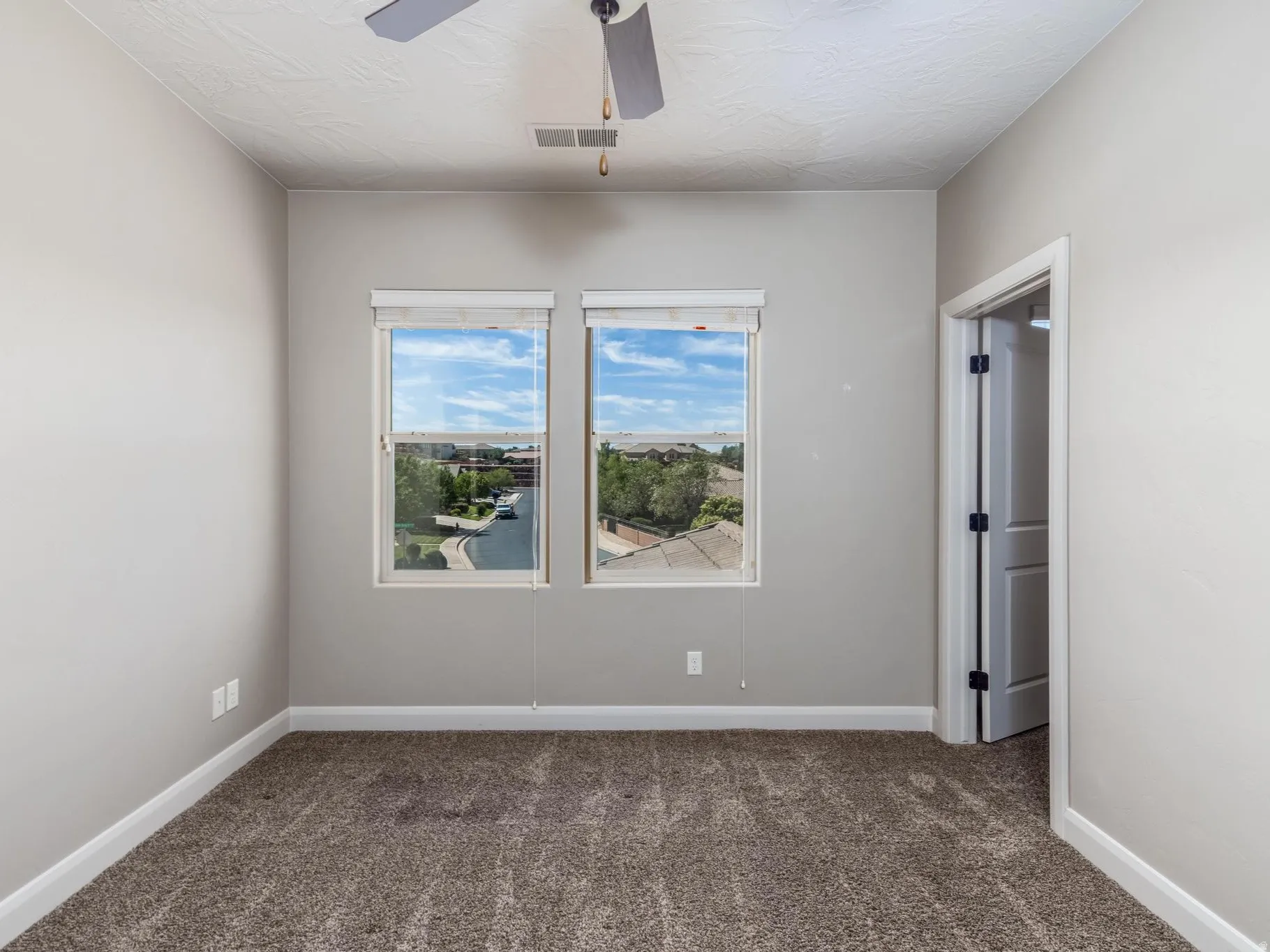 Dual windows with blinds, textured carpeting, neutral wall paint, white baseboards, and a ceiling fan with light fixture