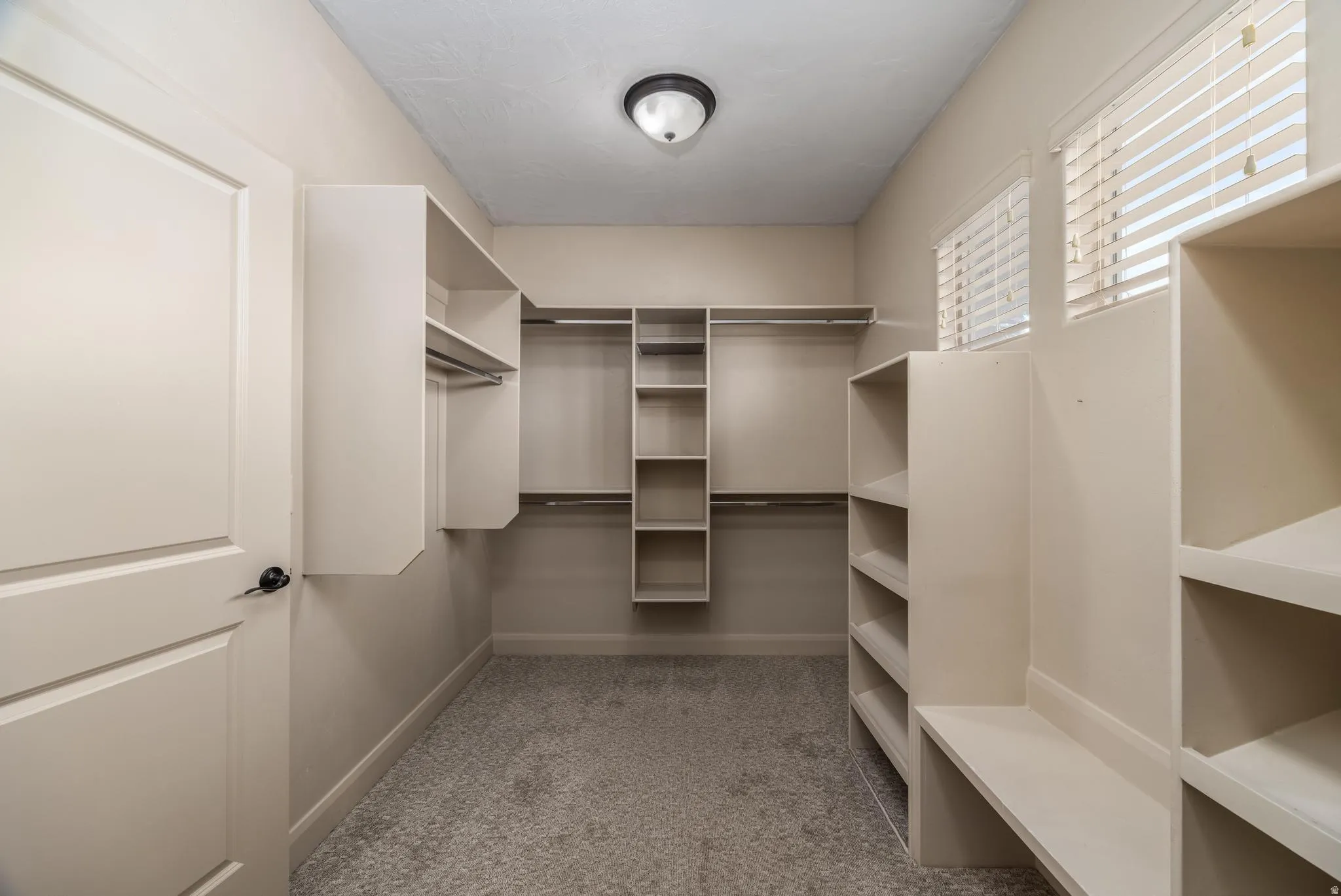 Walk-in closet featuring built-in shelving, hanging rods, two windows with blinds, and light gray carpet flooring