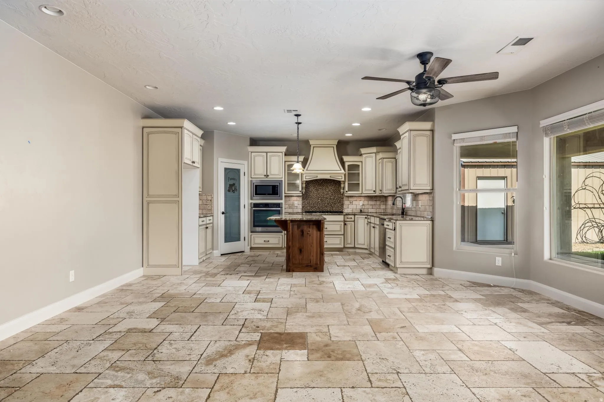 Spacious kitchen featuring stone tile flooring, cream-colored cabinetry, a wood-finish island, built-in double ovens, and a decorative range hood