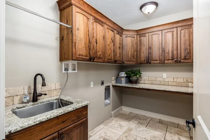 Laundry room featuring wood cabinetry, granite countertops, an undermount sink with a gooseneck faucet, a tile backsplash, and stone tile flooring