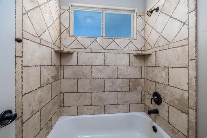 Bathtub and shower combination featuring neutral-toned tile surround with decorative accents, built-in corner shelves, a window with obscure glass, and oil-rubbed bronze fixtures