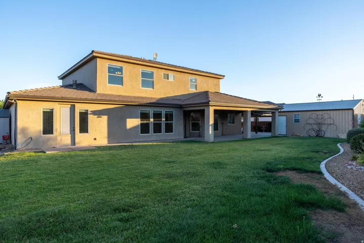 Expansive backyard featuring a large green lawn, covered patio, stucco exterior, and a detached storage building with a metal roof