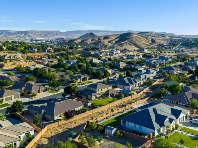 Suburban neighborhood featuring a variety of architectural styles and mature landscaping, framed by distant hills and a clear sky