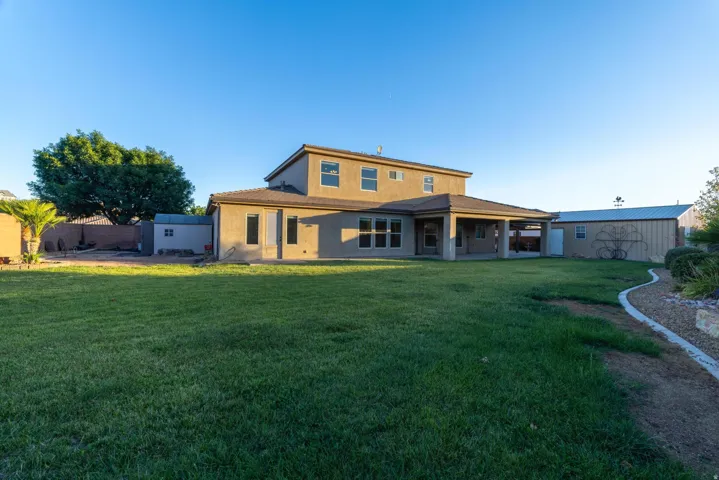 Expansive backyard featuring a large green lawn, covered patio, stucco exterior, and two visible storage sheds