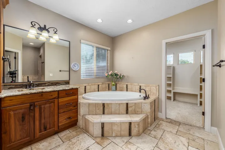 Spacious bathroom featuring a built-in jetted tub with tiled surround, a wood vanity with a granite countertop and undermount sink, and large format tile flooring