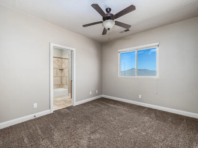 Carpeted room with a ceiling fan, a window with a roller shade, and an entryway to a bathroom featuring tiled walls and flooring