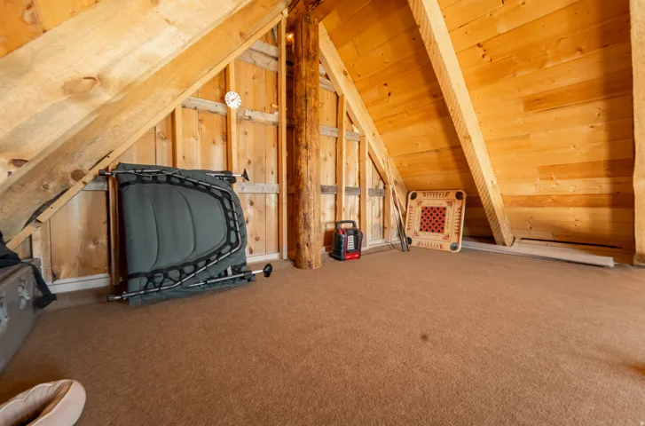 Expansive attic space featuring natural wood paneling, exposed wooden beams, a central wooden support column, and light brown carpeting