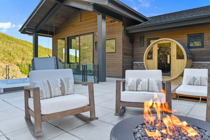Expansive outdoor patio featuring a fire pit with stone fill, a wood-sided barrel sauna, and a glass-paneled hot tub
