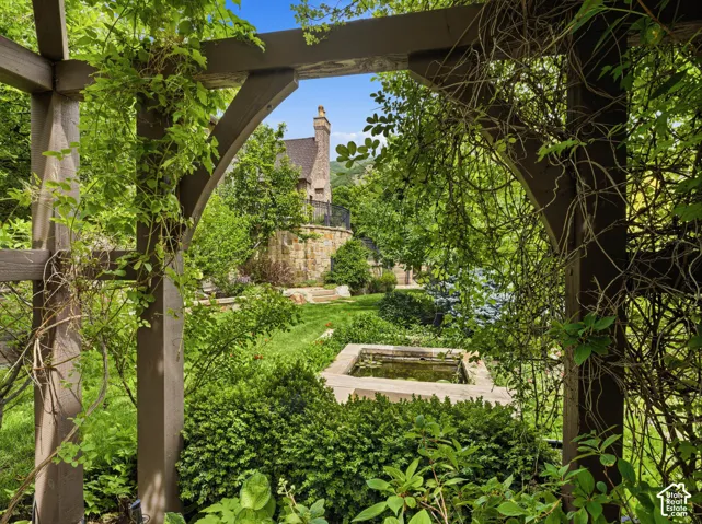 Lush backyard landscaping featuring a wooden pergola with climbing foliage, a central water feature, manicured lawns, mature trees, and a stone retaining wall