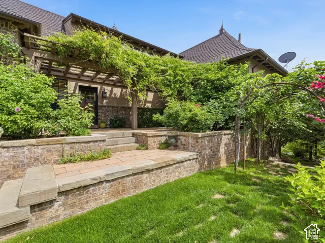 Terraced exterior landscaping featuring brick retaining walls with stone caps, stone paver patio, wood pergola with abundant vines, and a dark tile roof