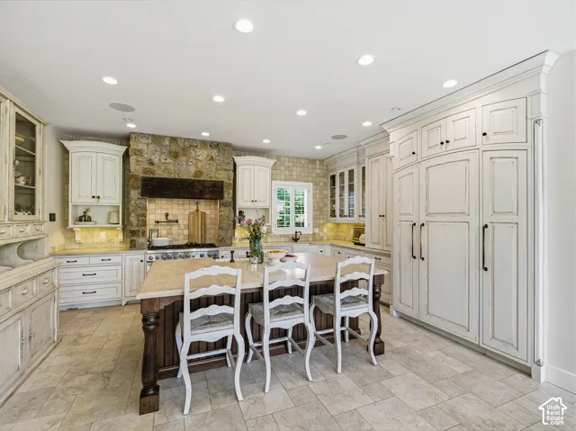 Spacious kitchen featuring a large central island with a light-toned countertop, extensive white cabinetry with dark hardware, a stone-clad range hood and backsplash, professional-grade range, and recessed lighting