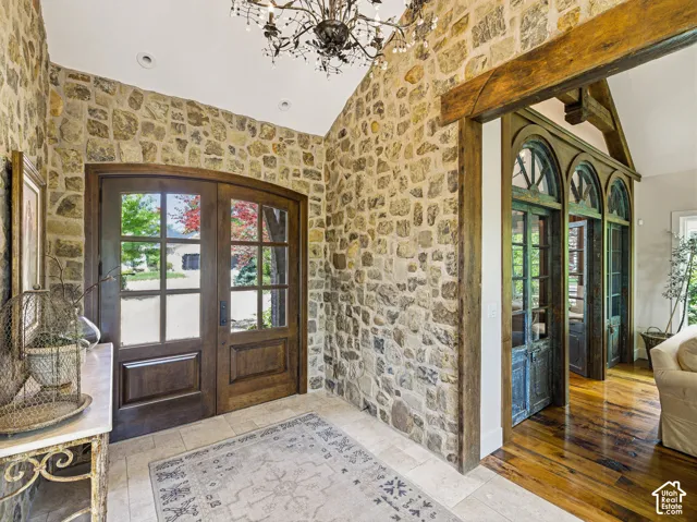 Foyer featuring stone walls, arched wood-frame glass panel double doors, a decorative chandelier, tile flooring, and a wood beam archway