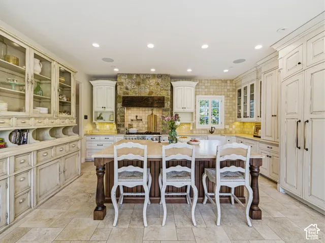Kitchen featuring a central island with a light-toned countertop, extensive built-in cabinetry with glass-front uppers, a stone-clad range hood, tile flooring, and recessed lighting