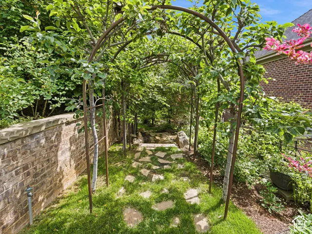 Landscaped garden pathway featuring a mature arbor with lush greenery, flagstone steps, manicured grass, a brick retaining wall, and verdant plantings