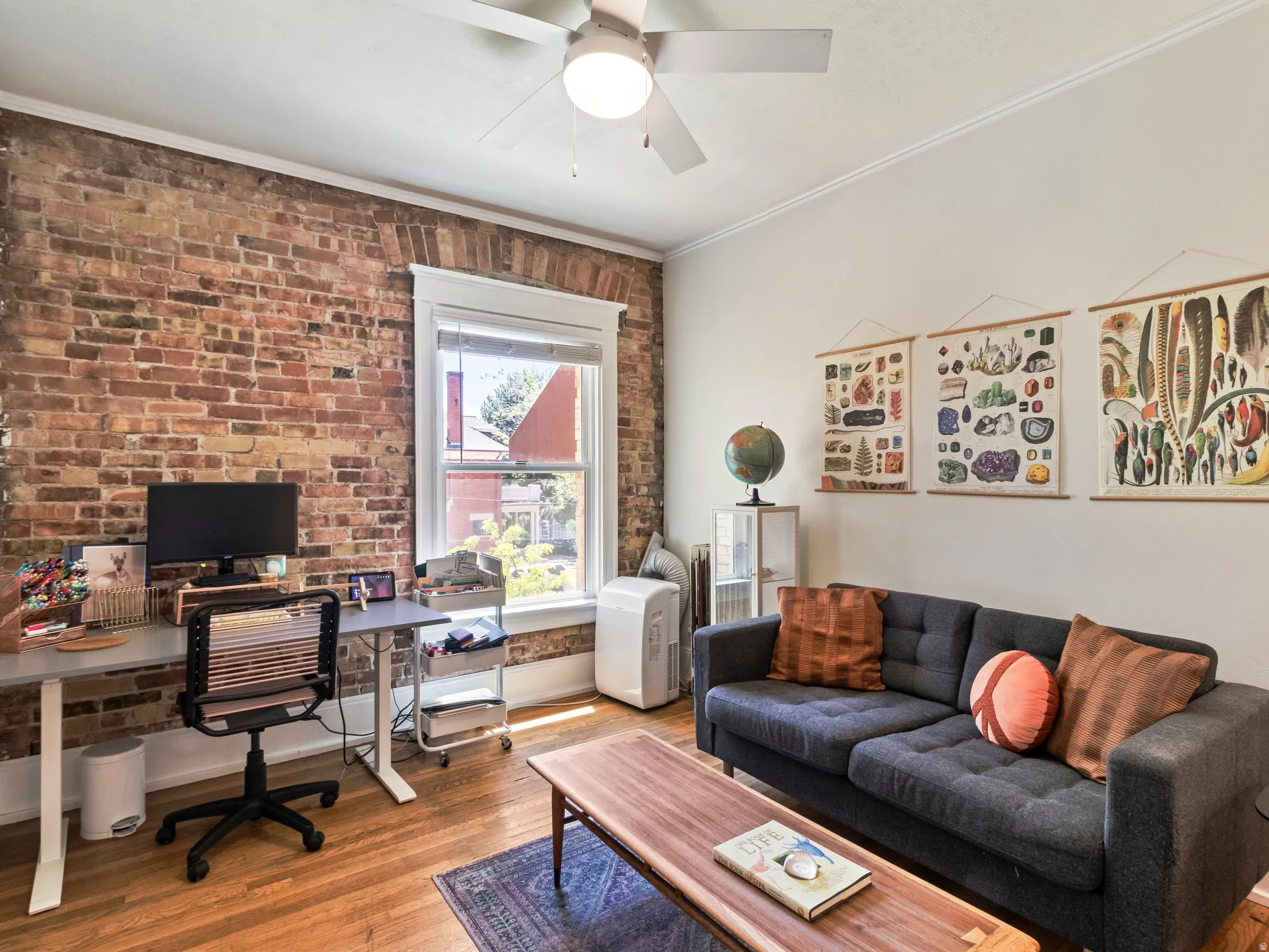 Exposed brick accent wall, wood-finish flooring, ceiling fan with integrated lighting, white trim window, and crisp white walls